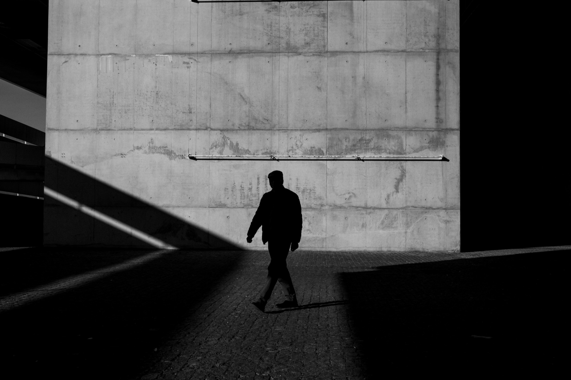 A lone figure walking past a vast concrete wall in deep shadow.